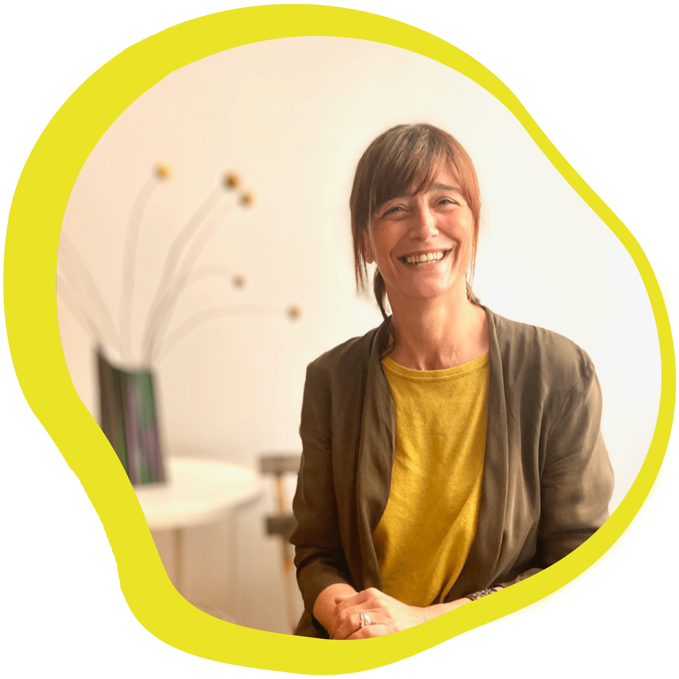 Una persona de cabello castaño, sonriente, con una camisa amarilla y una chaqueta marrón, está sentada en una habitación con una luz cálida. Sobre mí, hay un jarrón moderno con tallos altos decorativos en el fondo, enmarcado por un borde amarillo. Una persona de cabello castaño, sonriente, con una camisa amarilla y una chaqueta marrón, está sentada en una habitación con una luz cálida Sobre mí, hay un jarrón moderno con tallos altos decorativos en el fondo, enmarcado por un borde amarillo